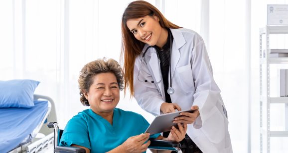 Asian medical doctor take care and explain senior elderly woman female patient on wheelchair with tablet, looking at camera